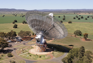 An aerial view of the Parkes radio telescope. The big dish is pointing away from and to the right. Several groups of people can be seen in the grounds around the telescope. In the distance is a mountain range.