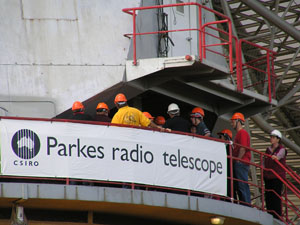A group of people in orange hardhats are on the azimuth track of the Parkes telescope behind a banner with the CSIRO logo and 'Parkes radio telescope' on it