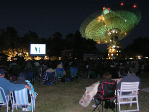 In the foreground many people on blankets and picnic chairs watch the movie with the telescope in the background