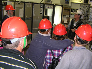 Brett in a white hard hat (back right) is facing a group of people in red hard hats (their backs to camera). There is various electronic equipment in the background.