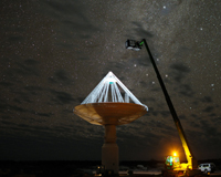 A long exposure image of a white dish-like ASKAP antenna at night, with a green receiver at the top of the antenna, and a tall telehandler next the antenna. Credit: Alex Cherney/terrastro.com.