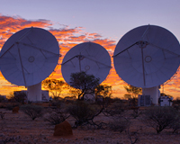 Three white dish-like ASKAP antennas sit amongst red dirt and desert bushes in the foreground of a desert sunset. Credit: Alex Cherney/terrastro.
