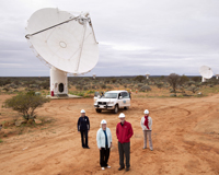 Four people looking at the camera in front of several dish-like antennas in an Australian outback setting