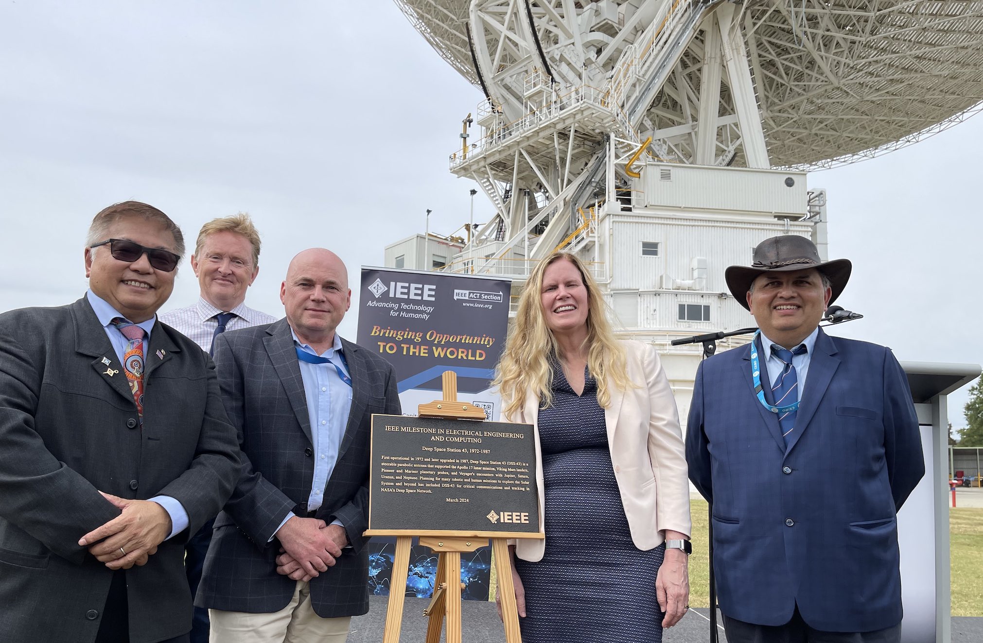 A group of people around the IEEE plaque, with the Tidbinbilla 70m antenna in the background.