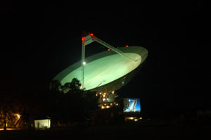 The Parkes radio telescope lit up at night with a movie screen in the front showing the dish in the movie the dish pointing in a similar direction