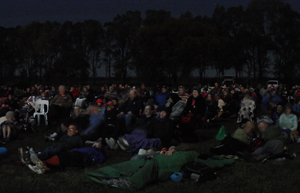 A crowd of people at night are sitting on blankets and picnic chairs. They are all looking off left where the movie is screening