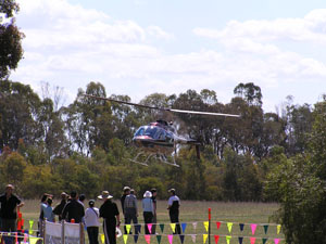 A helicopter hovers just above the ground. A group of people stand just beyond some bunting. Trees are in the background.