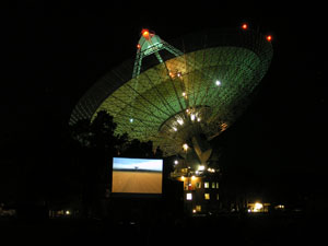 An aerial shot with wheatfields in the foreground appears on a movie screen with the telescope behind