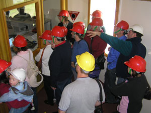 A group of people in red and yellow hard hats look through a large window (left) into a room of computers. Lewis in a white hard hat (back right)  is behind the group explaining the computers.