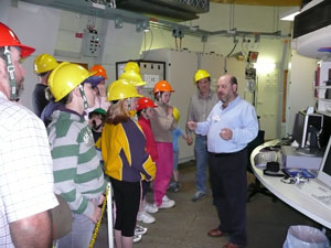 John Sarkissian at the right is facing a large group of people (left) in yellow hard hats who are listening to him. There is various electronic equipment behind them.