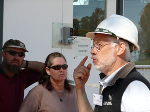Dr Dave McConnell in a white hard hat points to his eye. There are two people in the background who are part of the larger group (off left of frame)