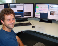 A male (Tim Shimwell) in the foreground of two computer screens showing calculations and maps.